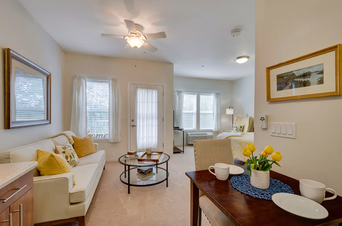 A bright and cozy assisted living apartment featuring a beige sofa with yellow and floral cushions, a round glass coffee table, a small dining table set with white dishes and a vase of yellow tulips, and a bed with a yellow floral comforter near windows with white curtains. The room has light-colored walls, carpeted floors, a ceiling fan, and framed artwork on the walls.