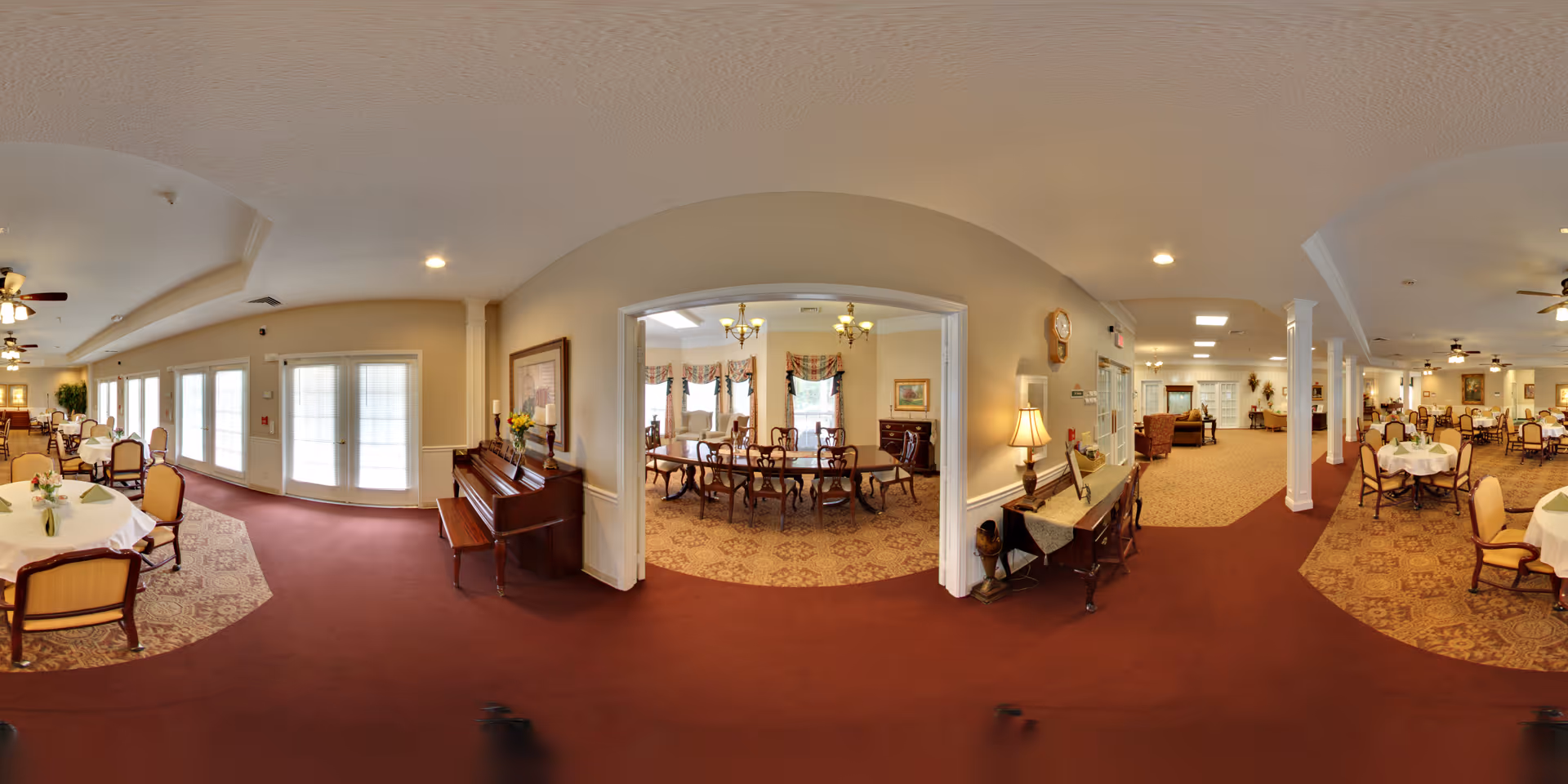 A spacious senior living facility dining area with multiple round tables covered with white tablecloths and surrounded by cushioned chairs. The room features large windows allowing natural light, ceiling fans, and a piano against one wall. Adjacent to the dining area is a smaller room with a long wooden dining table and chairs, decorated with framed artwork and floral curtains. The carpet is a mix of red and patterned designs, and the walls are painted in neutral tones with decorative molding.