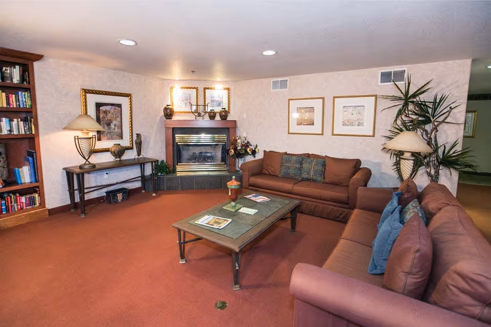 A cozy living room with two brown sofas adorned with decorative pillows, a rectangular coffee table with magazines and a decorative item, a fireplace with a wooden mantel, framed artwork on the walls, a tall indoor plant, a bookshelf filled with books, and two table lamps providing warm lighting.