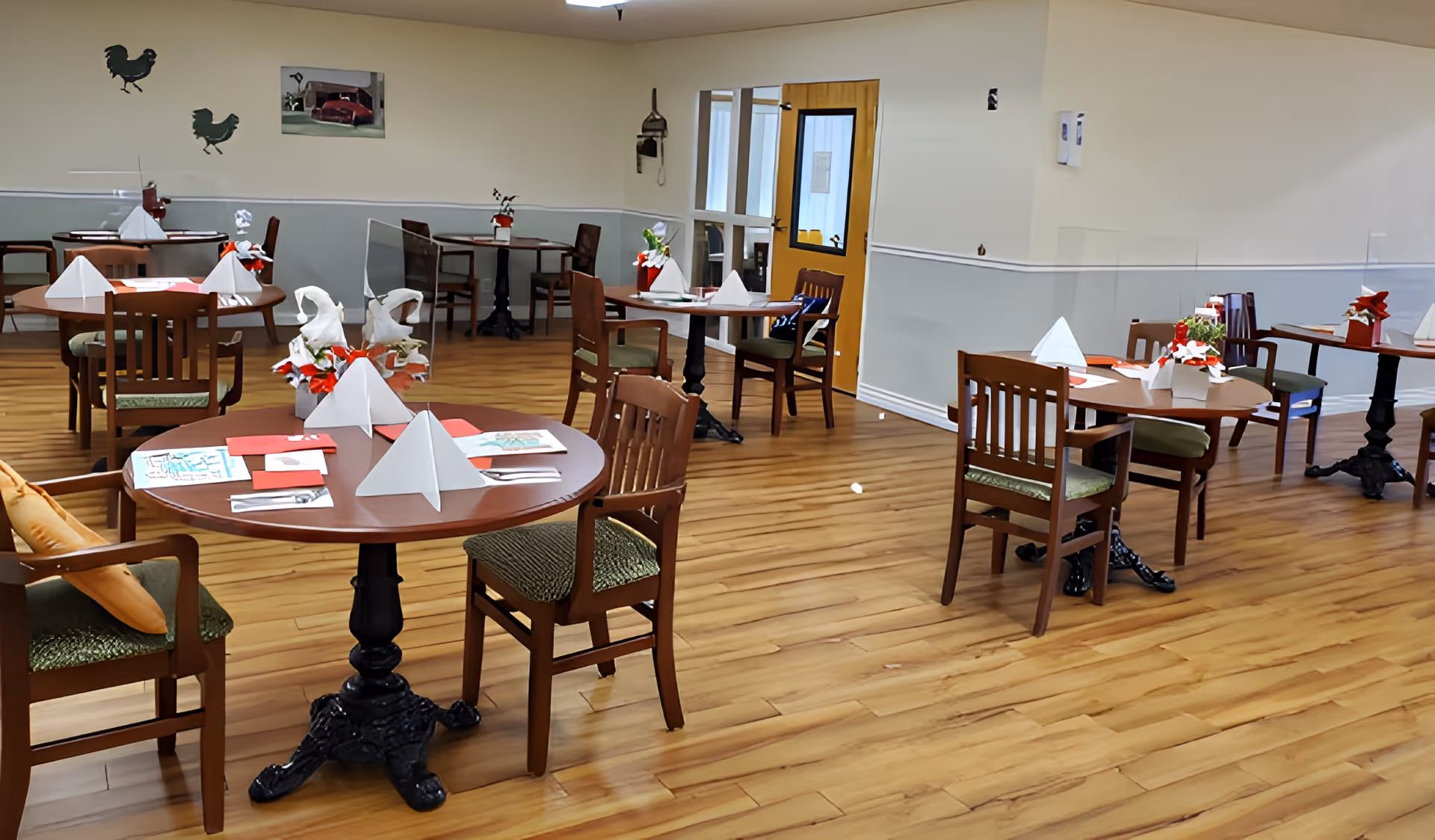 Dining room with round tables and chairs set with napkins and floral centerpieces on a wood floor.