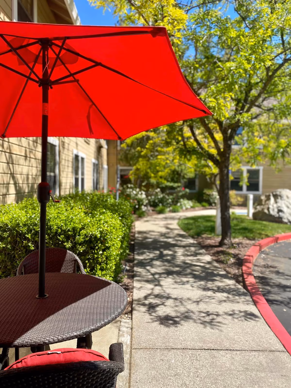 Outdoor patio area at Hilltop Commons Senior Living featuring a round wicker table with a red umbrella and matching red cushioned chairs. The patio is adjacent to a building with beige siding and white-framed windows. A concrete walkway curves through a landscaped garden with green bushes, trees, and flowering plants under a clear blue sky.