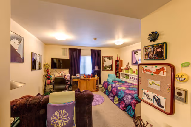 A brightly decorated assisted living bedroom with a single bed, sofa, TV, and colorful wall art.