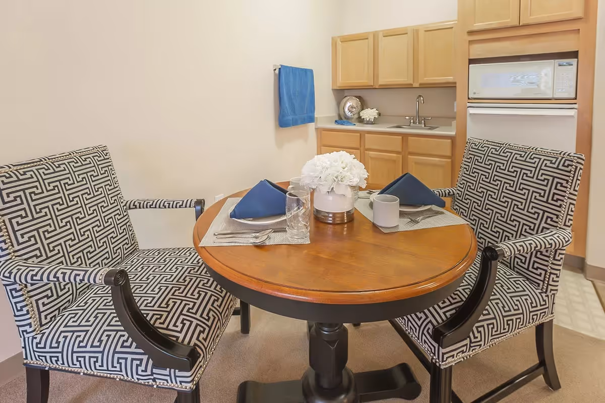 A small dining area with a round wooden table set for two with blue napkins, glassware, and a white flower centerpiece. Two patterned armchairs with black and white geometric designs are positioned around the table. In the background, there is a kitchenette with light wood cabinets, a sink, a microwave, and a blue towel hanging on the wall.