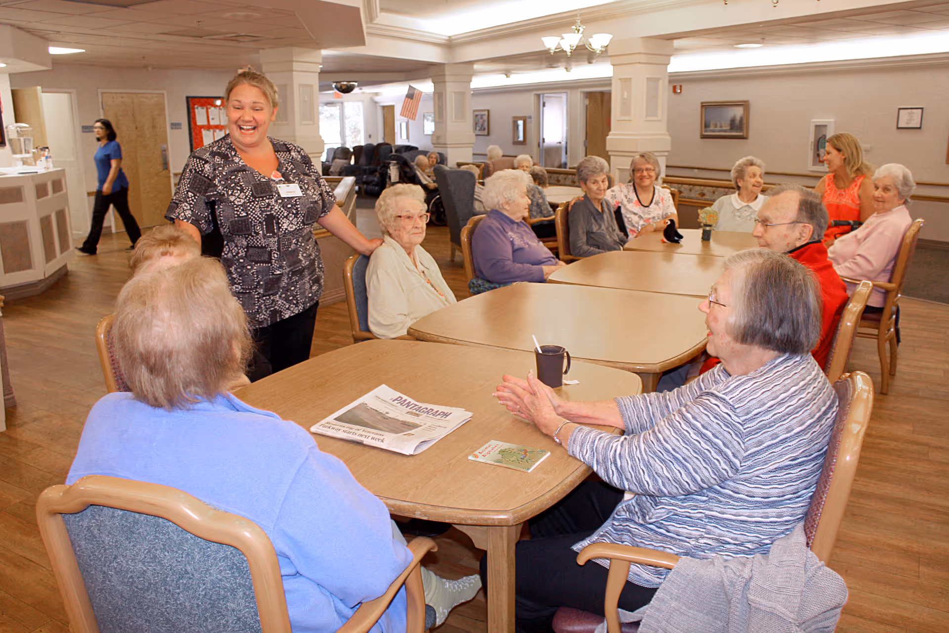 A smiling staff member talks with a group of elderly residents seated around tables in a bright communal dining/activity room.