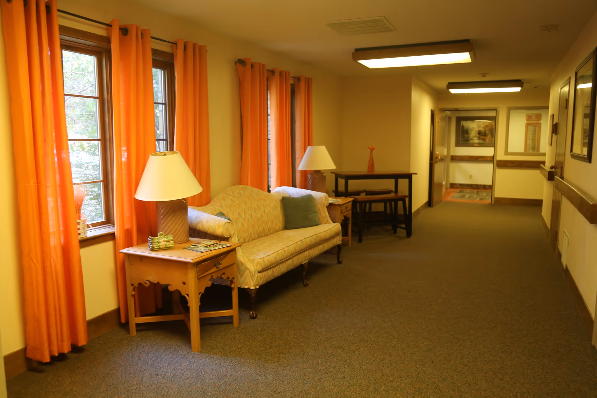 A hallway in a senior living facility with orange curtains on windows to the left, a patterned loveseat with green pillows, two wooden side tables each with a lamp, and a small table with two stools further down the hall. The walls are light yellow and the floor is carpeted.
