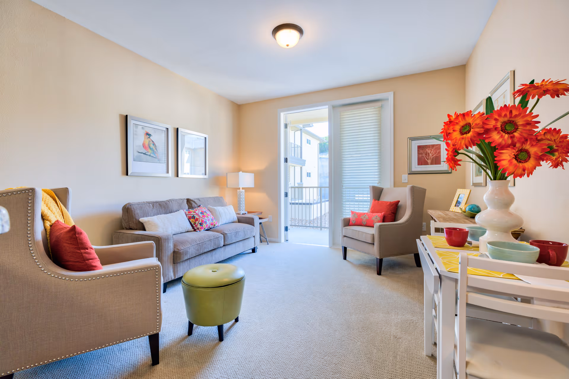 A bright and cozy living room with beige walls and carpet. The room features a gray sofa with colorful pillows, two beige armchairs with red cushions, a small green ottoman, a side table with a lamp, and a white table with bowls and a vase of vibrant orange flowers. There are framed pictures on the walls and a glass door leading to a balcony or outdoor area.