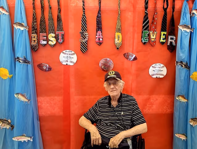 An elderly man wearing a striped black shirt and a black cap sits in a wheelchair in front of a red wall decorated with hanging neckties spelling out 'BEST DAD EVER'. The wall also features cutouts of baseballs, footballs, and basketballs, with blue curtains on either side adorned with fish decorations.
