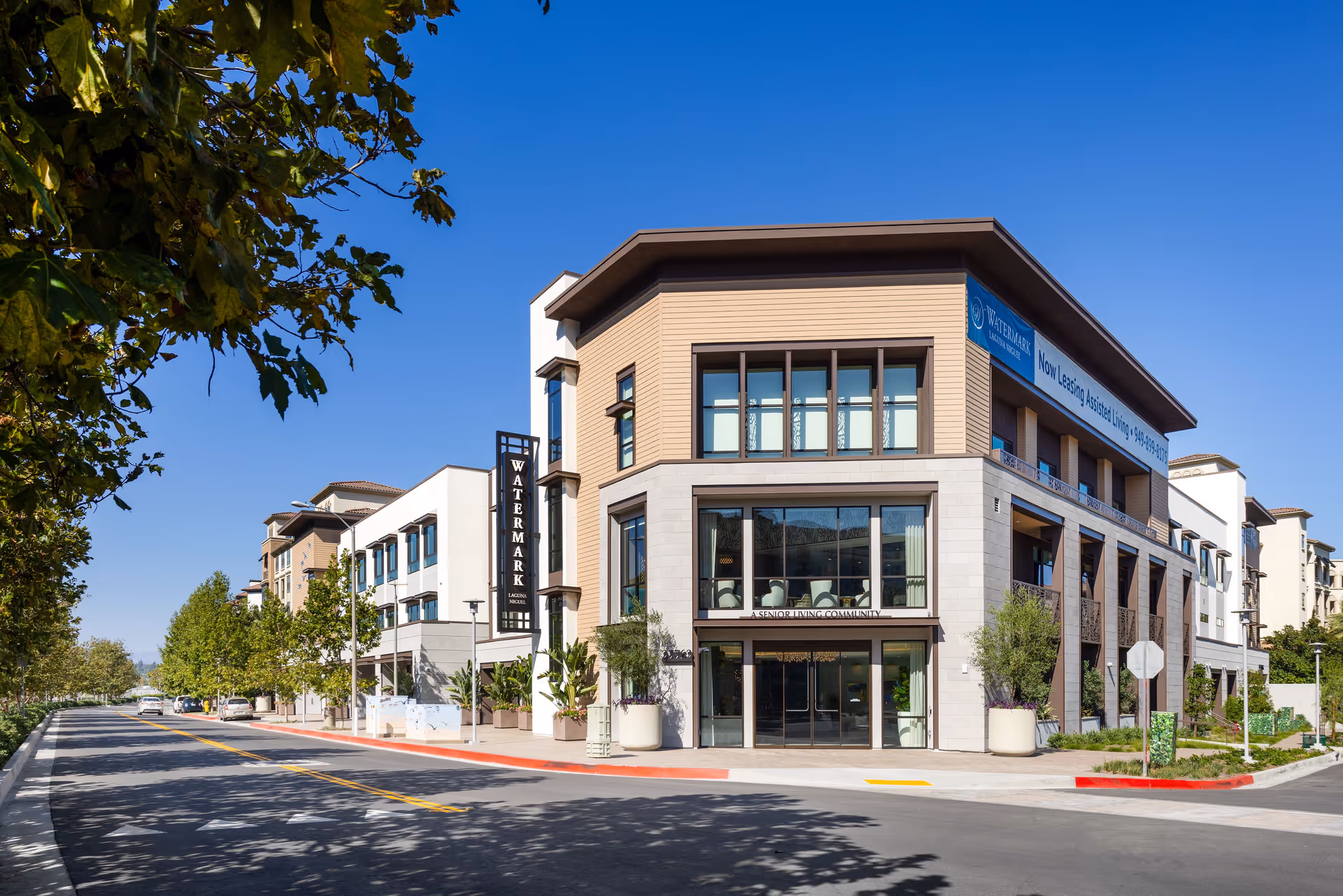 Exterior view of Watermark Laguna Niguel, a senior living community building, on a sunny day with clear blue sky. The building has large windows, modern architectural design, and a sign displaying the facility name. Trees line the street in front of the building.