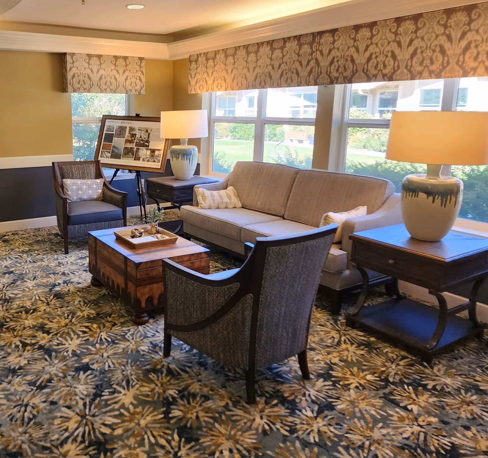 A cozy senior living facility lounge area with a beige sofa, two armchairs, a wooden coffee table, and two side tables each holding a decorative lamp. Large windows with patterned valances let in natural light, and a framed display board is positioned near the corner.