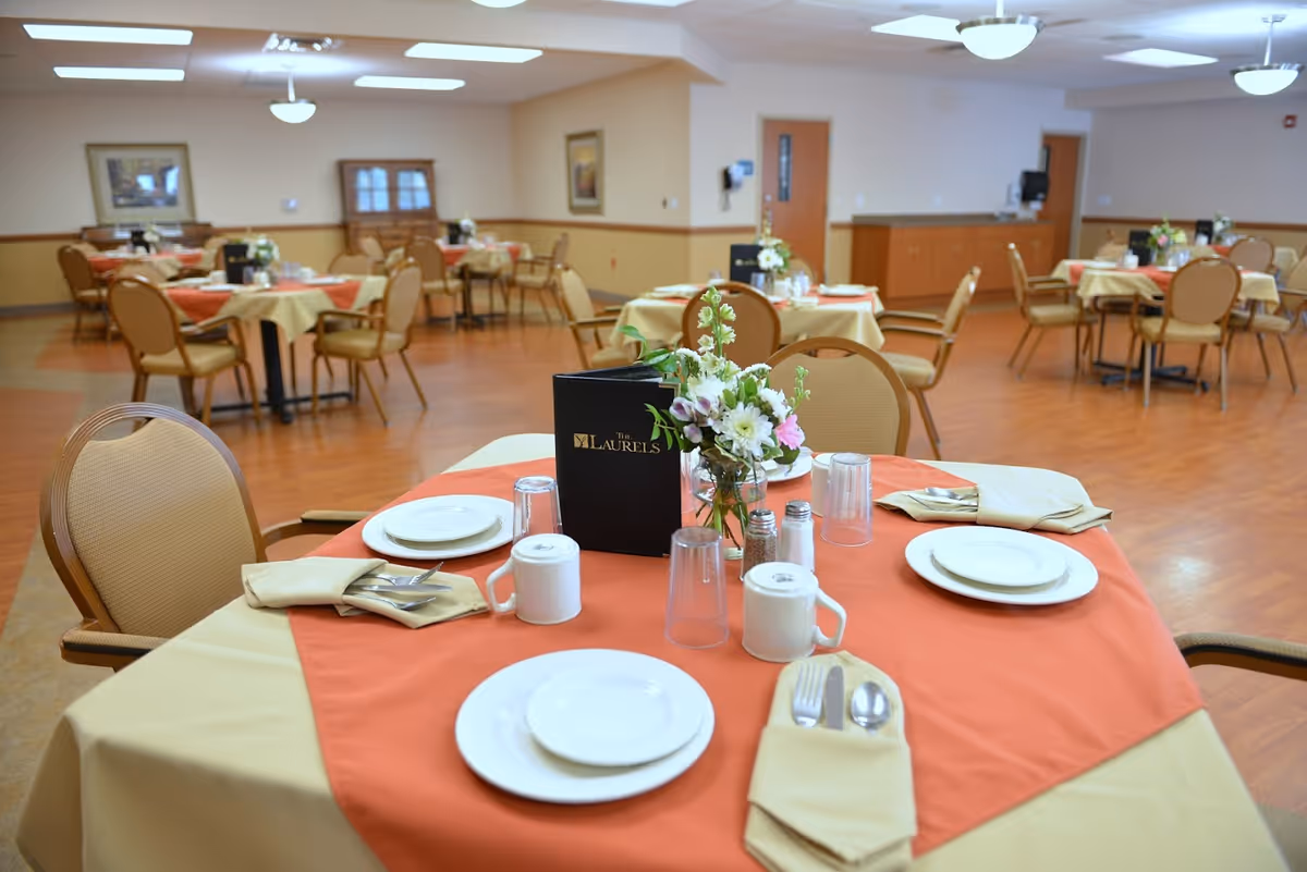 A dining room with multiple round tables covered with beige and orange tablecloths. Each table is set with white plates, silverware wrapped in beige napkins, cups, and a small flower arrangement. The room has wooden flooring, beige walls with a wooden trim, and ceiling lights. A menu labeled 'The Laurels' is placed on the table in the foreground.