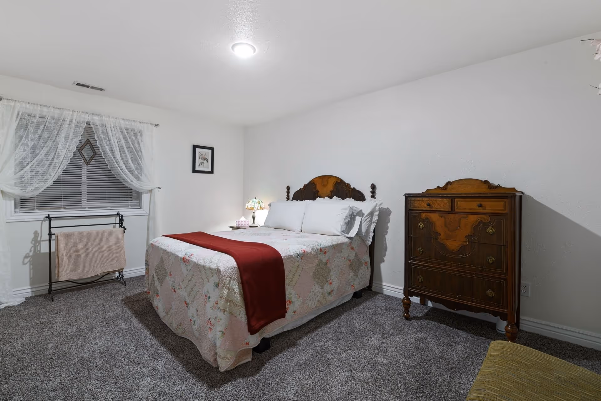 A cozy bedroom with a vintage wooden bed featuring a floral quilt and a red throw blanket. There is a wooden dresser with ornate details on the right side and a small nightstand with a decorative lamp and tissue box next to the bed. The room has white walls, a window with lace curtains, and a carpeted floor.