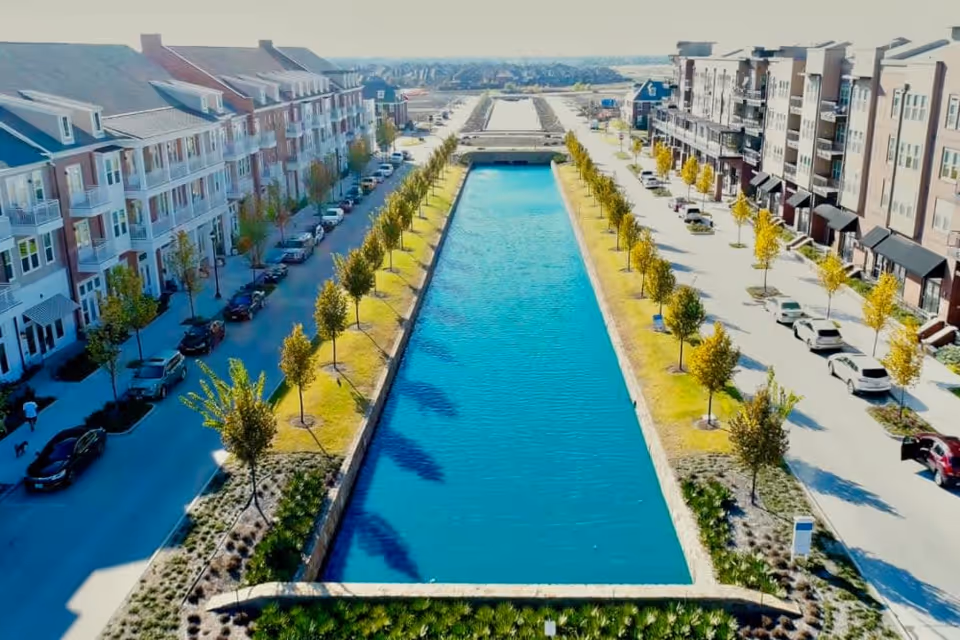 Aerial view of a long, narrow water canal flanked by rows of young trees and parked cars on both sides, with multi-story residential buildings lining the streets in a suburban neighborhood.