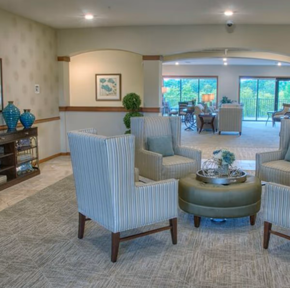 A spacious and well-lit living room area with four striped armchairs arranged around a round ottoman with a decorative tray and flowers. In the background, there are large windows and sliding glass doors that offer a view of greenery outside. The room features neutral tones, carpeted floors, and decorative elements including blue vases on a wooden console table and framed artwork on the wall.