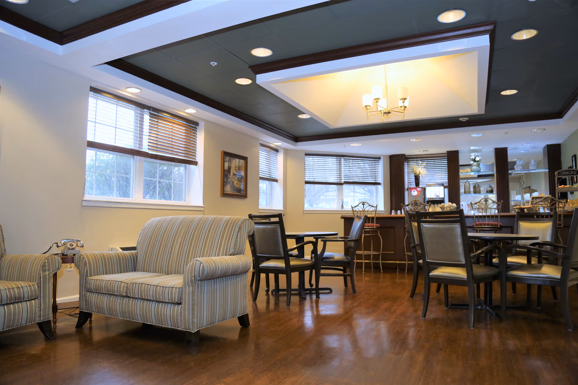 A cozy senior living common area with striped upholstered armchairs, several round tables with chairs, large windows with wooden blinds, and a counter area with bar stools and a chandelier overhead.