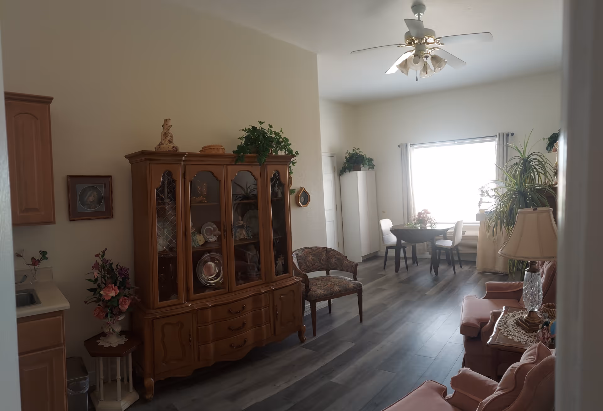 A bright living room area with wooden flooring, featuring a wooden china cabinet filled with decorative plates and items, a floral upholstered chair, a small round dining table with two chairs near a large window, several potted plants, and a pink upholstered sofa with a side table and lamp.