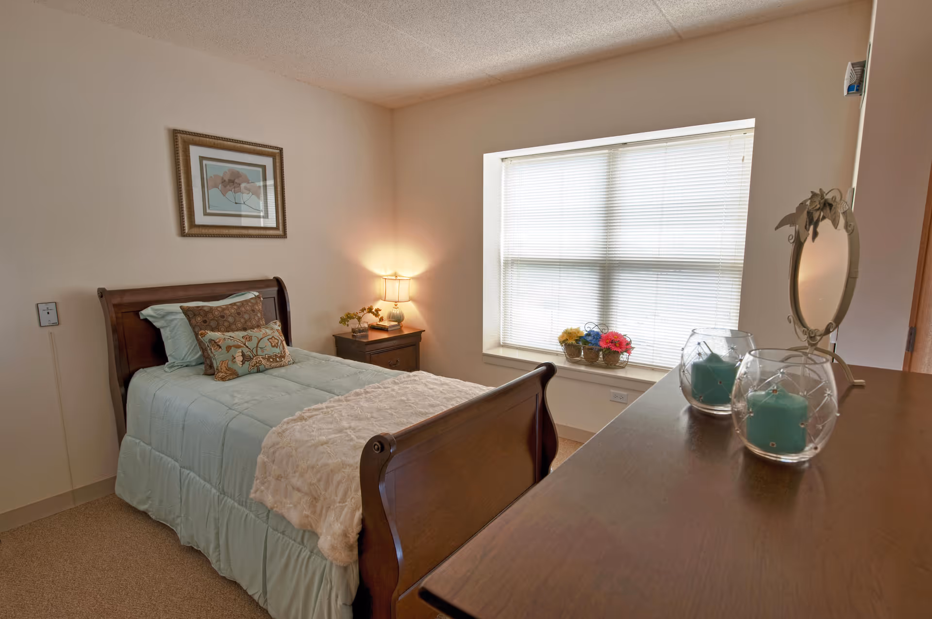 Sunlit bedroom with a wooden sleigh bed, nightstand, window with blinds and a dresser topped with decorative candles and a mirror.