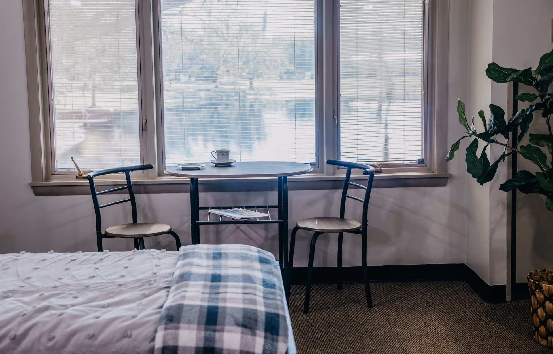 Bed with a plaid blanket in the foreground, a small round table and two chairs by large windows overlooking water, and a potted plant.