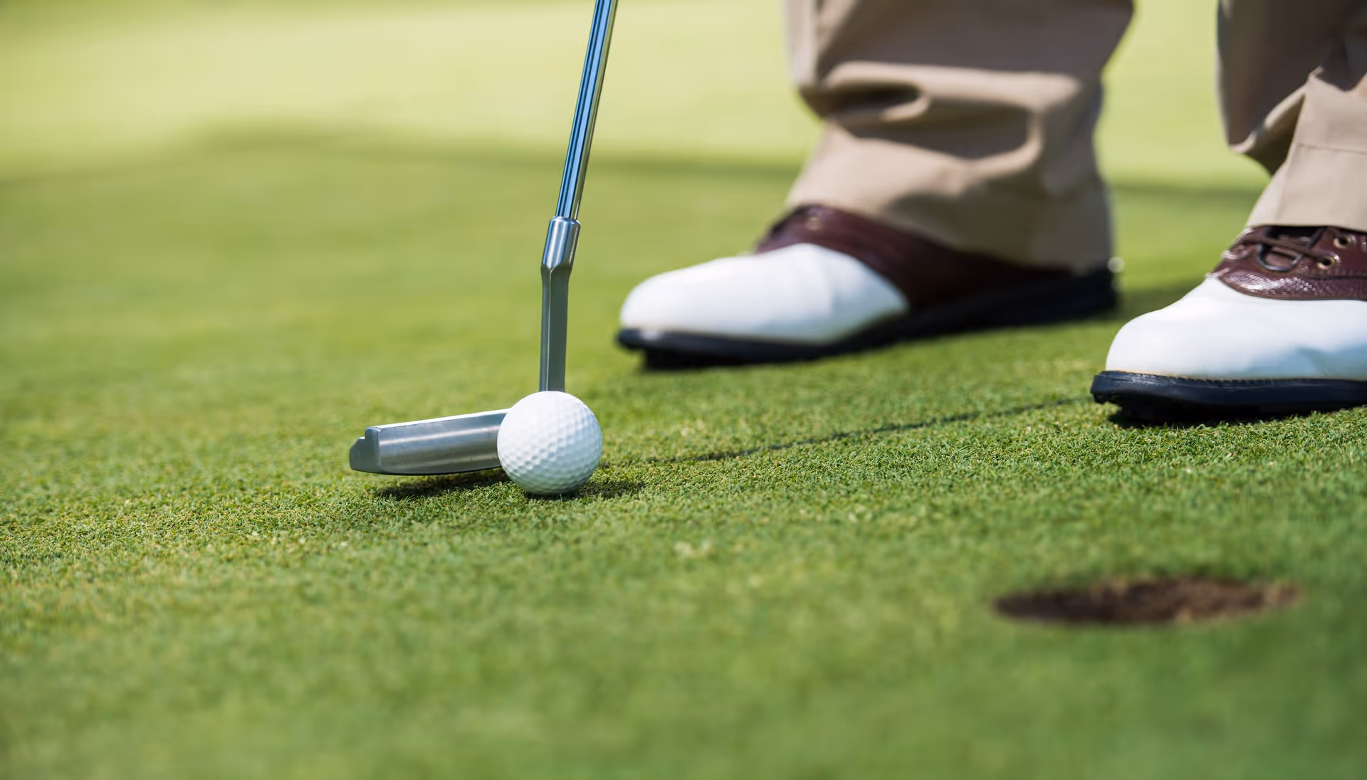 Close-up of a golf putter and ball on a putting green next to a golfer's shoes.
