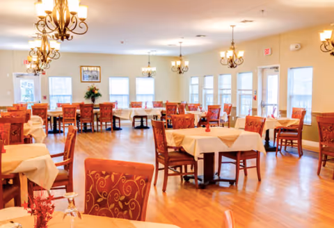 A spacious dining room with multiple tables covered in beige tablecloths and surrounded by red cushioned chairs with a gold swirl pattern. The room has wooden flooring, several chandeliers hanging from the ceiling, large windows letting in natural light, and a few floral decorations on the tables and in the corner.