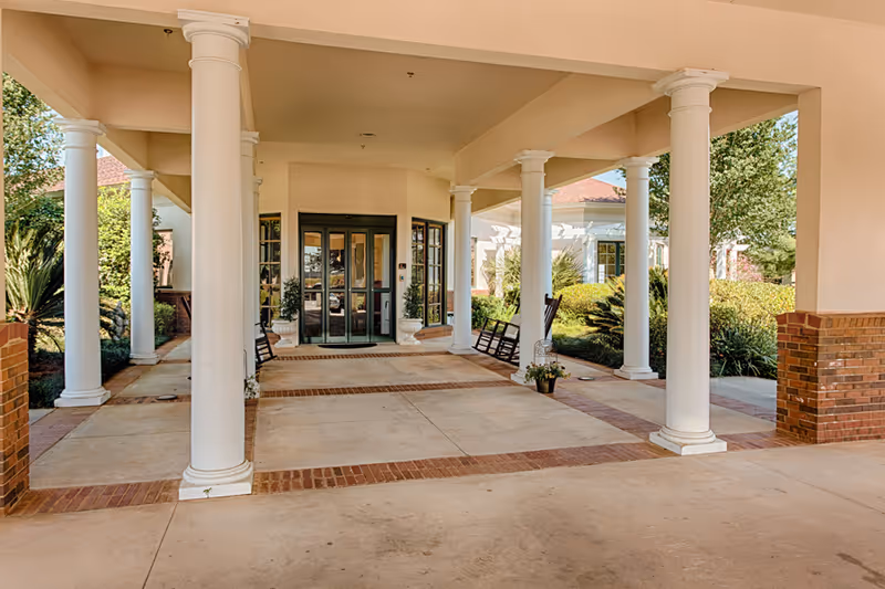 Covered entrance area of a building with white columns and brick accents, featuring glass double doors at the far end and rocking chairs on either side. There are plants and greenery visible around the entrance.