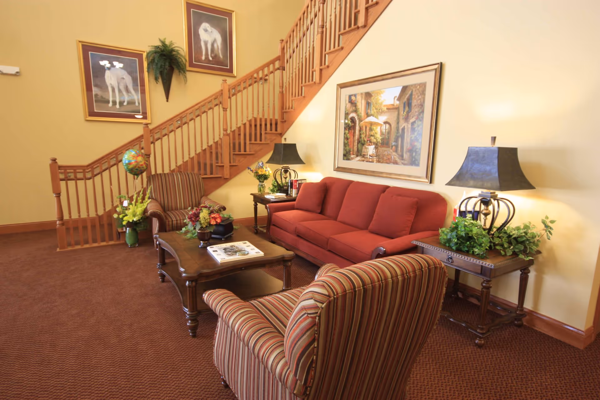 A cozy living room area with a red sofa, two striped armchairs, a wooden coffee table with books and flowers, two side tables with lamps and plants, and a wooden staircase in the background. The walls are decorated with framed paintings and a wall-mounted plant.