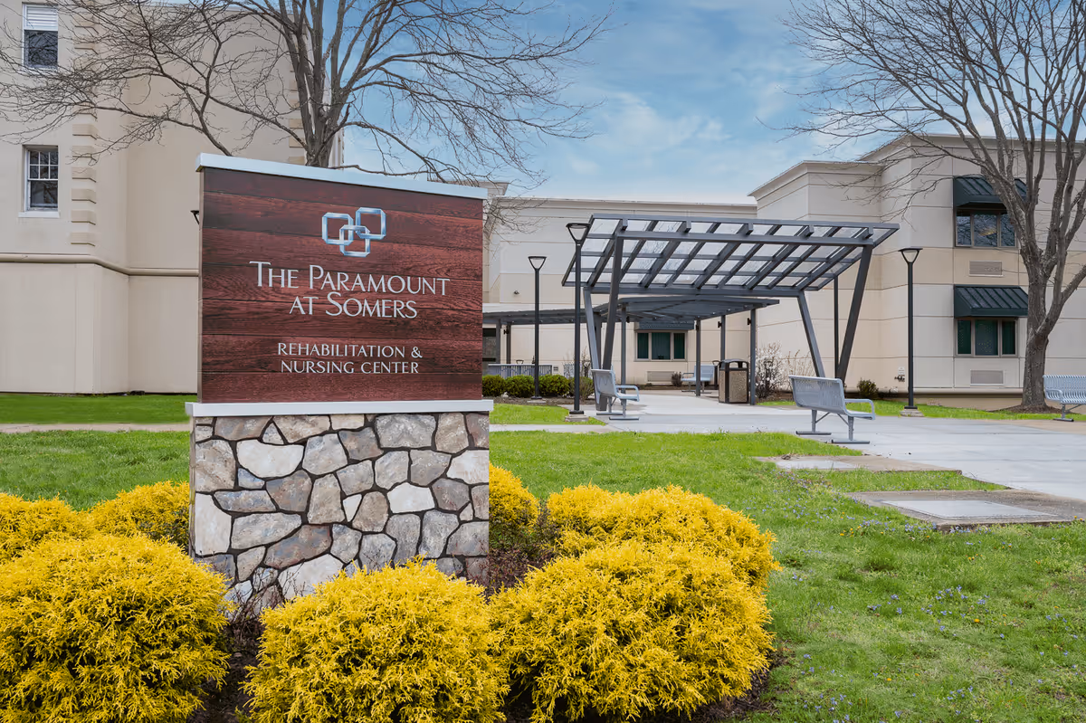 Outdoor view of The Paramount at Somers Rehabilitation & Nursing Center showing a stone and wood sign with the facility name, surrounded by yellow bushes and green grass. In the background, there is a building with windows, leafless trees, and a covered seating area with benches.
