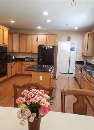 A spacious kitchen with wooden cabinets and hardwood floors. The kitchen features a black refrigerator, a built-in oven, a stovetop on a central island, and a double sink. In the foreground, there is a table with a vase of pink and white flowers. The room is well-lit with recessed ceiling lights.