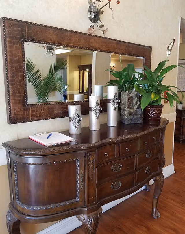 A wooden sideboard with ornate carvings and claw feet, topped with decorative candles, a potted plant, and a rock sculpture. Above the sideboard is a large rectangular mirror with a textured brown frame. The reflection in the mirror shows part of the room with another plant and furniture.
