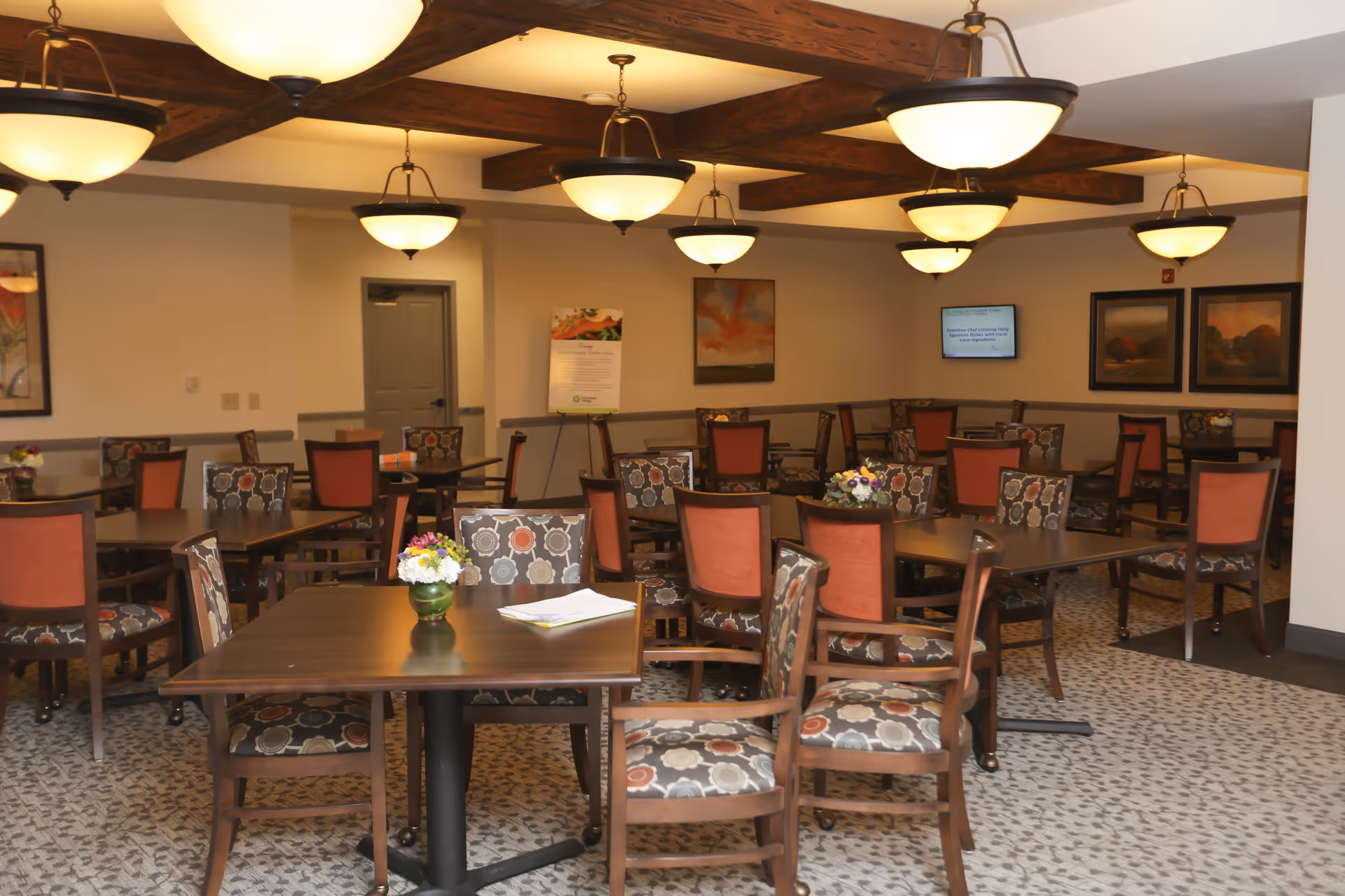 Well-lit dining room with multiple tables and patterned chairs, ceiling lights, and small floral centerpieces.