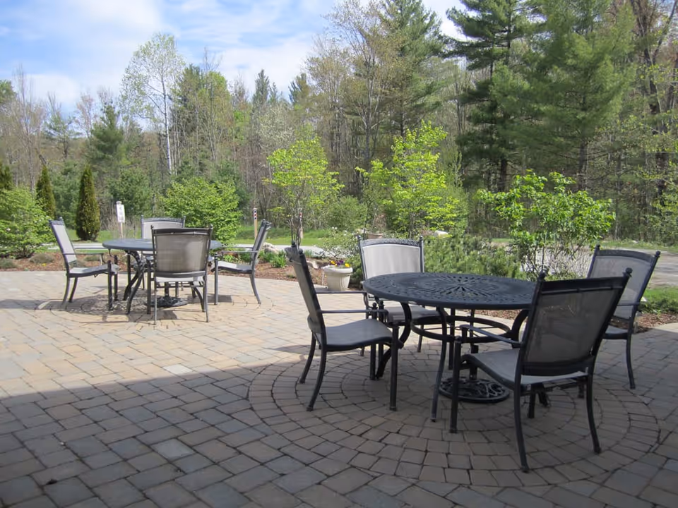 Outdoor patio area with two round metal tables and several metal chairs with mesh backs and seats, surrounded by greenery and trees under a partly cloudy sky.