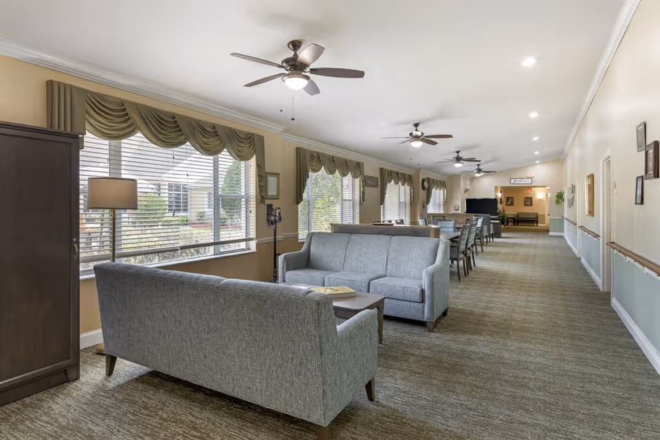 A long, well-lit common area with multiple seating arrangements including two gray sofas around a coffee table and several dining tables with chairs. The room has large windows with green valances, ceiling fans with lights, and framed pictures on the walls. The carpet is patterned in neutral tones and the walls are painted beige with white trim.