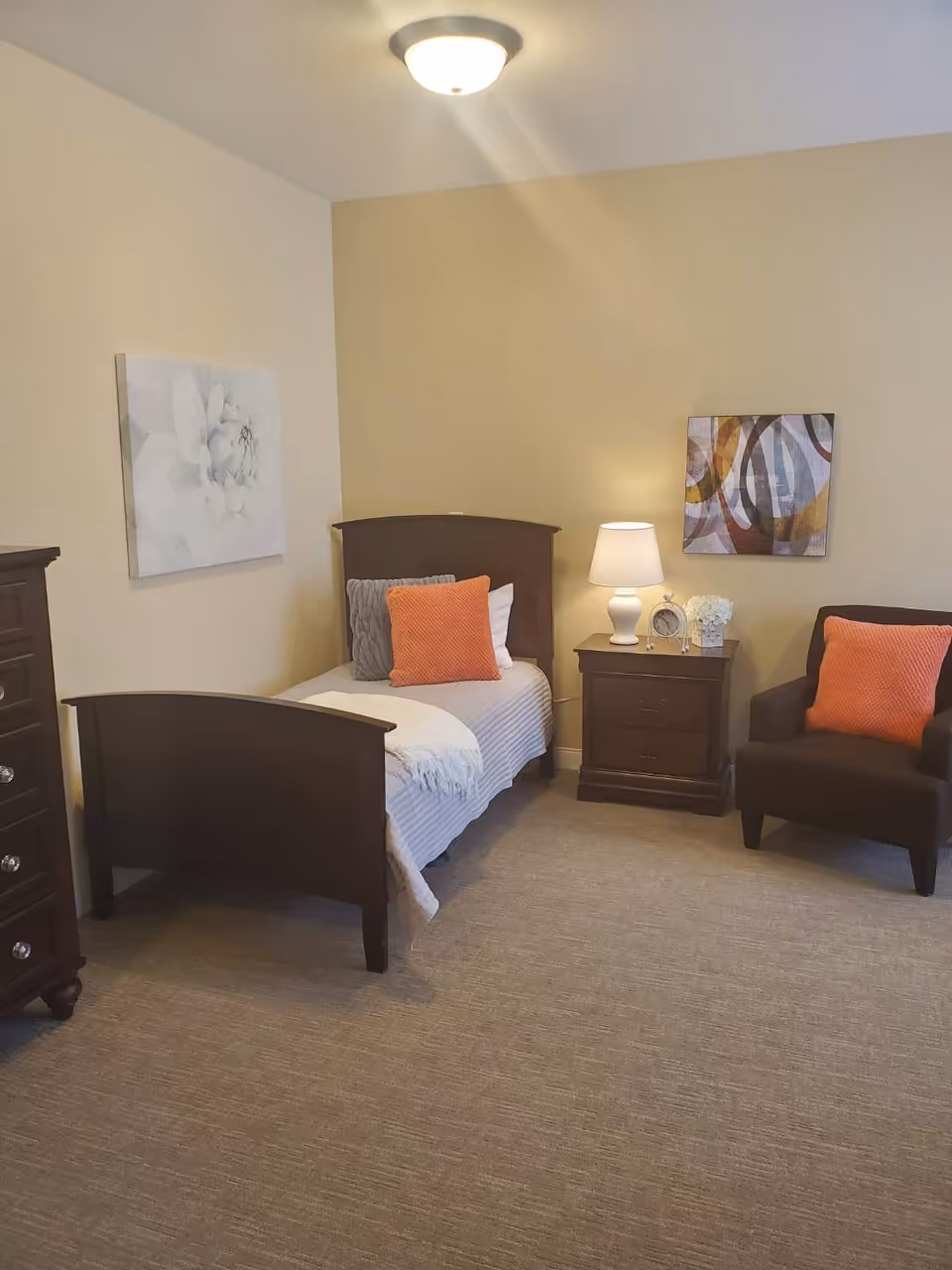 A cozy bedroom with a single wooden bed featuring gray and orange pillows and a white blanket. Next to the bed is a wooden nightstand with a white lamp, a clock, and a small flower arrangement. There is a dark brown armchair with an orange pillow to the right of the nightstand. The walls are painted beige and decorated with two pieces of artwork. The floor is carpeted in a neutral tone.