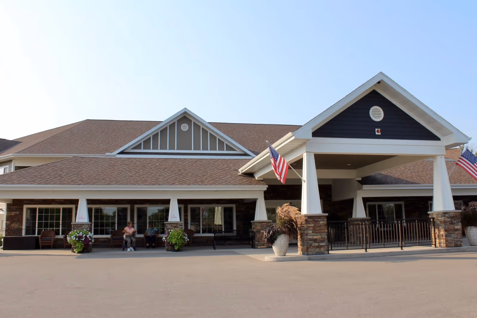 Front exterior view of a senior living facility named Bay Harbor of La Crescent, featuring a covered entrance with two American flags, brick pillars, large windows, and a few people sitting on chairs outside.
