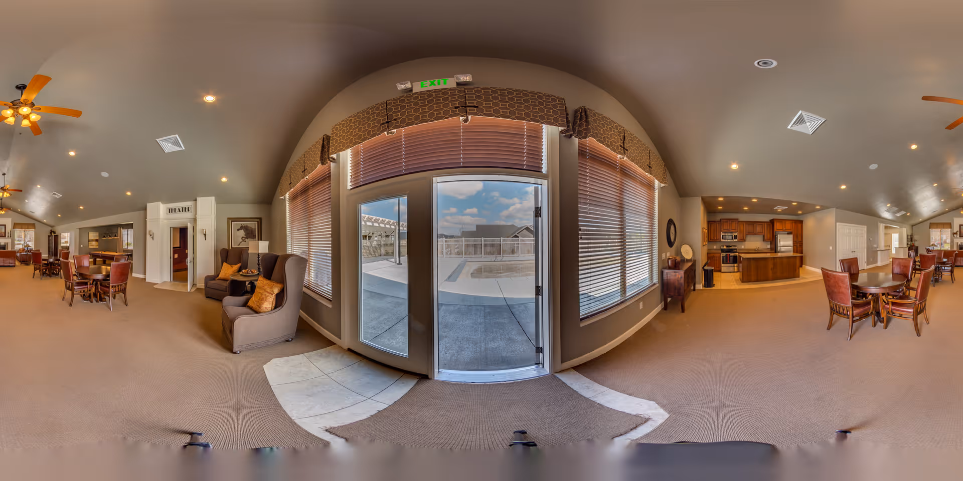 Wide panoramic view of a senior living facility common area featuring multiple seating arrangements with tables and chairs, a cozy sitting area with armchairs and cushions, ceiling fans, and a kitchen area with wooden cabinets and appliances. Large windows and a glass door provide a view of an outdoor patio and pool area under a partly cloudy sky.