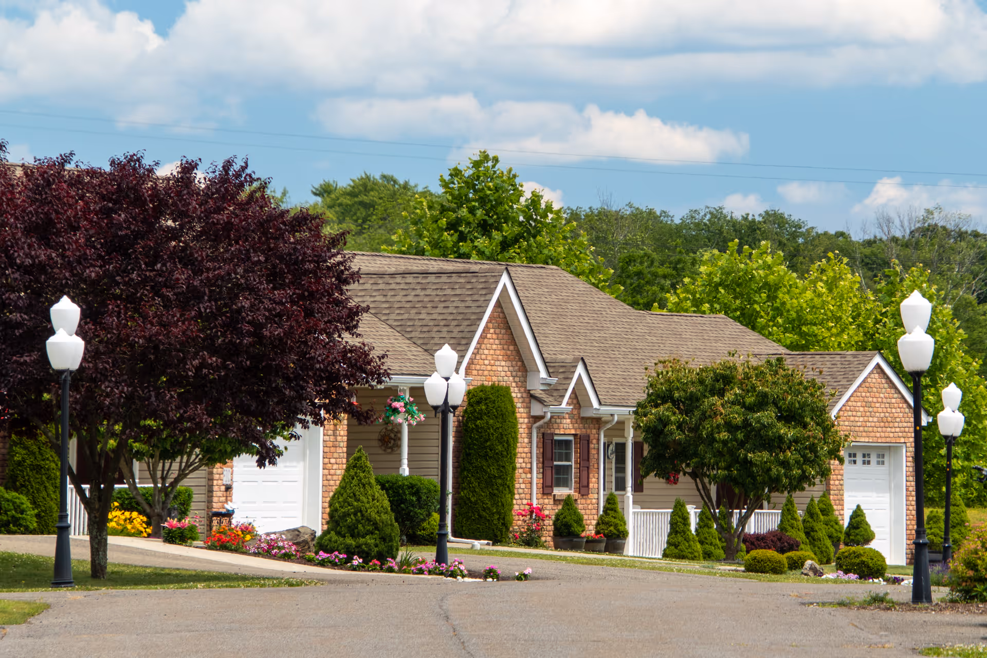 Brick-faced single-story residential building with garages, manicured shrubs, lamp posts, and flowering beds under a partly cloudy sky.