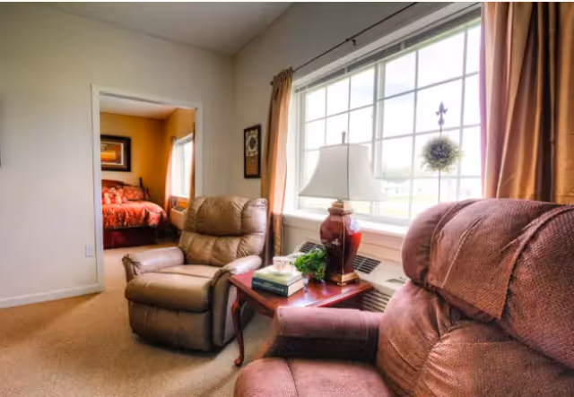 A cozy living area with two recliner chairs, a wooden side table with a lamp, books, and a small plant. A large window with curtains lets in natural light. In the background, a bedroom with a bed covered in a red patterned bedspread is visible through an open doorway.