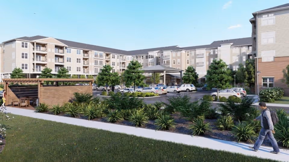 Exterior view of a multi-story senior living facility with beige and brick facade, surrounded by landscaped greenery and a parking lot with several cars. A person is walking on the sidewalk in the foreground, and there is a wooden pergola structure with seating on the left side.
