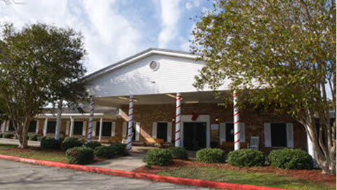 Exterior view of a single-story nursing and rehabilitation facility with a covered entrance supported by columns wrapped in red and white stripes, surrounded by bushes and trees under a partly cloudy sky.