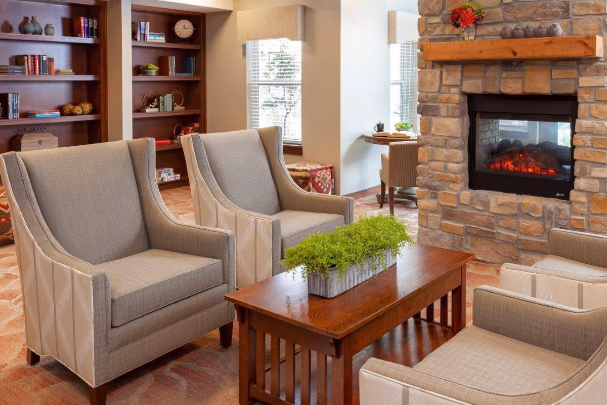 A cozy living room area featuring four beige upholstered armchairs arranged around a wooden coffee table with a rectangular planter of green plants. Behind the chairs is a stone fireplace with a wooden mantel decorated with a small red flower arrangement. To the left, there are built-in wooden bookshelves with books and decorative items. A window with white blinds allows natural light into the room, and a small desk with a chair is visible in the background.