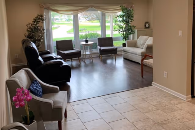 Bright common living area with multiple chairs and sofas arranged by large windows overlooking a green lawn.