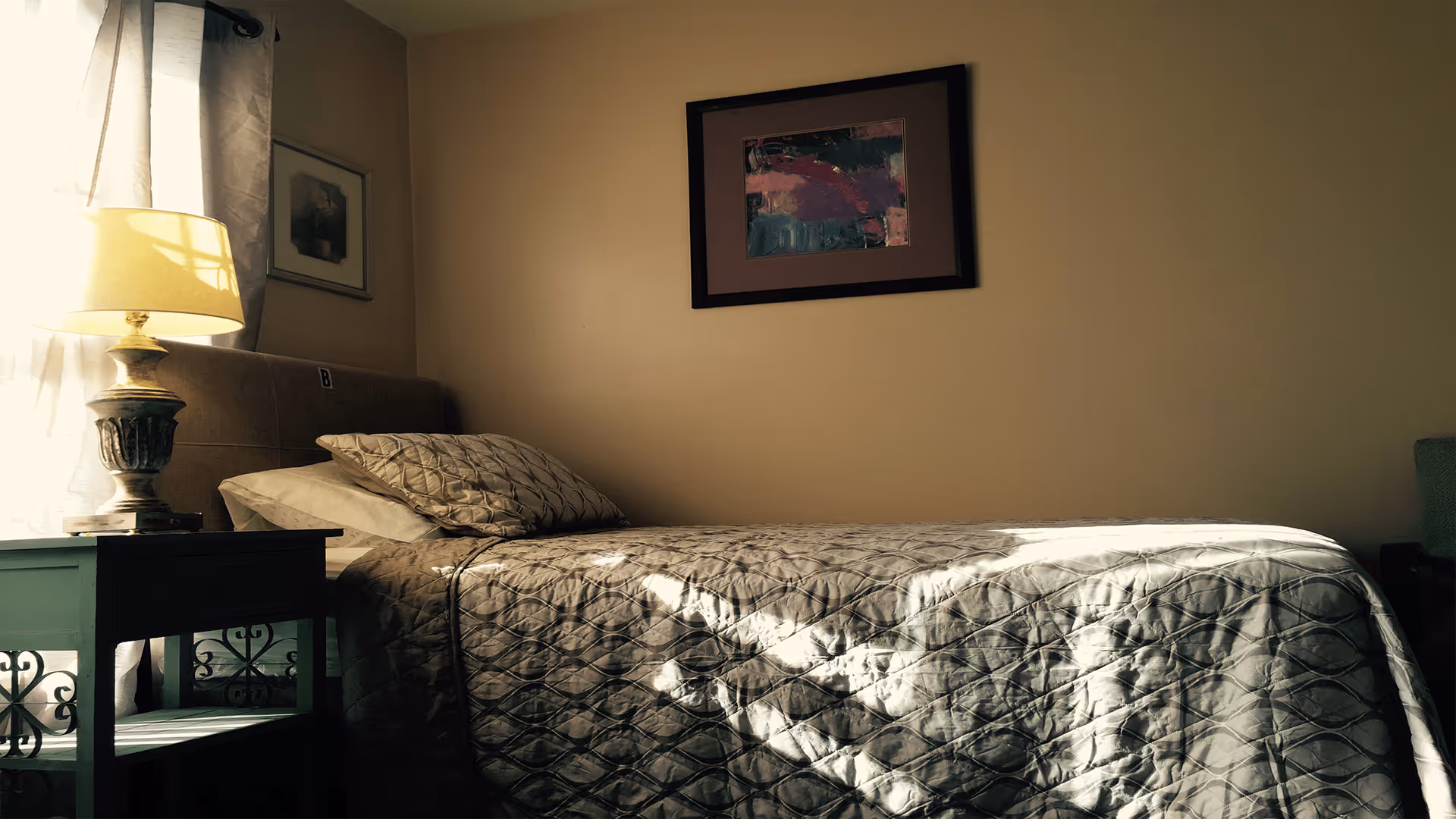 A single bed with a patterned quilt and two pillows in a softly lit room. Next to the bed is a green nightstand with a decorative lamp. There are two framed pictures on the beige walls, one above the bed and one near the window with sheer curtains.