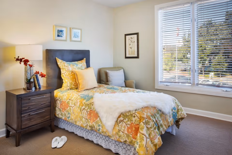 A cozy bedroom with a single bed covered in a floral patterned yellow, orange, and green bedspread, topped with multiple pillows and a white fur throw. Next to the bed is a wooden nightstand with a lamp, a small potted plant with red flowers, and two books. A pair of white slippers is placed on the carpeted floor beside the nightstand. There is a comfortable armchair in the corner and a large window with white blinds letting in natural light. The walls are light-colored with two framed pictures hanging.