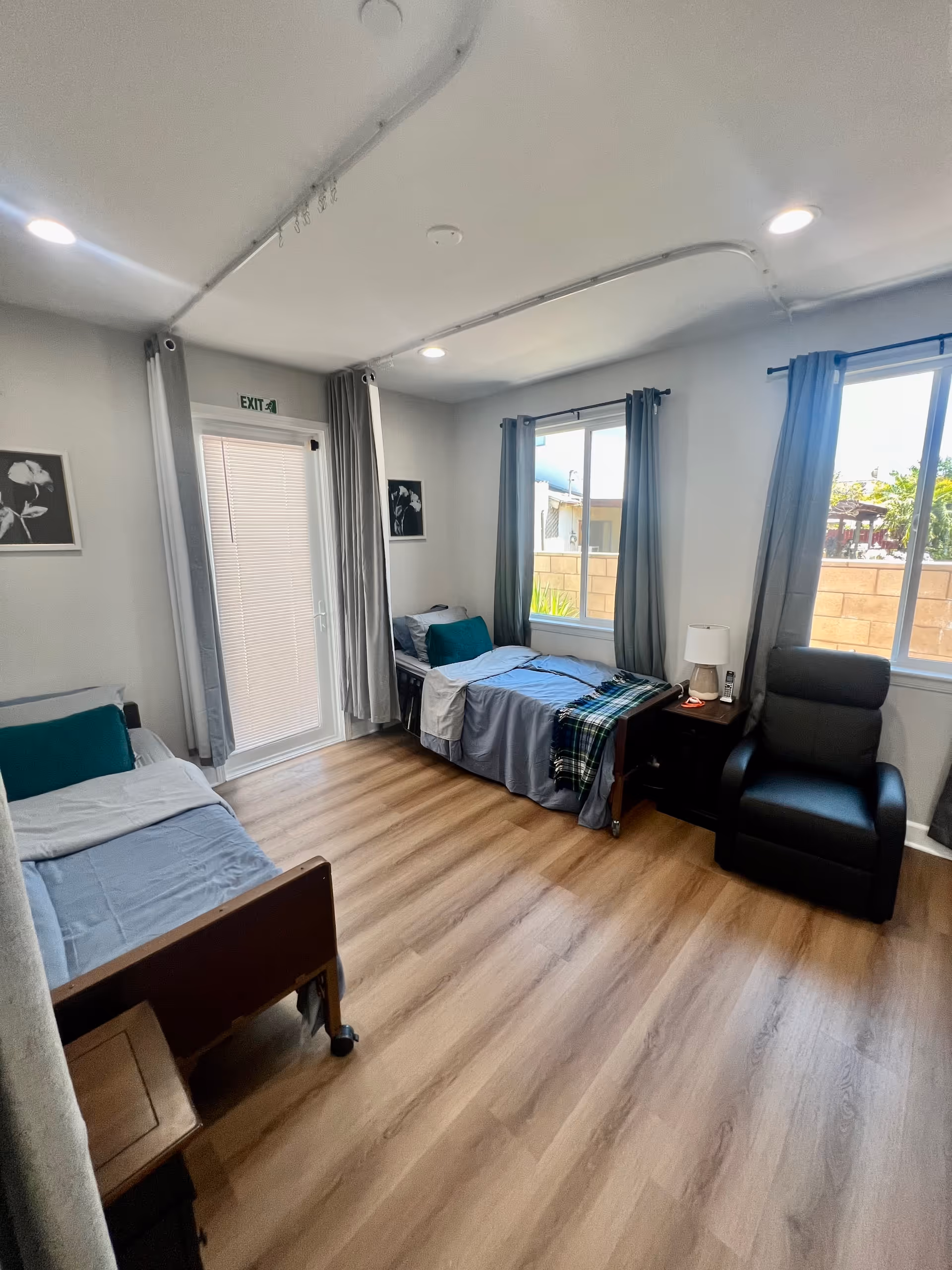 A bright and clean bedroom with two single beds, each with blue bedding and teal pillows. There is a black armchair near two windows with gray curtains. A small wooden nightstand with a lamp and a phone sits between one bed and the armchair. The room has light wood flooring and white walls with two black and white floral pictures. An exit door with a window and blinds is visible on the left side.