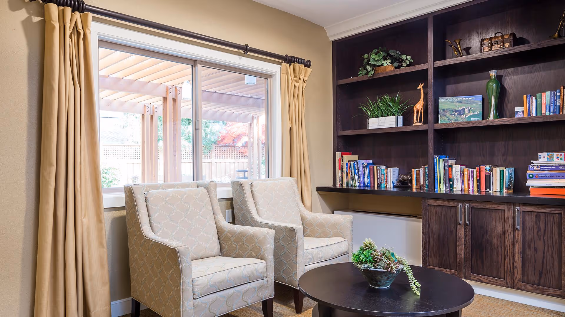 A cozy sitting area with two beige patterned armchairs facing a round black coffee table with a small plant centerpiece. Behind the chairs is a large window with beige curtains, showing an outdoor pergola and garden. To the right, there is a dark wooden built-in bookshelf filled with books and decorative items.