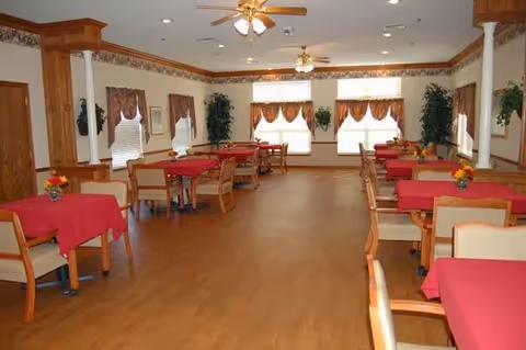 A spacious dining room with multiple wooden tables covered with red tablecloths and surrounded by beige cushioned chairs. The room has wooden flooring, ceiling fans with lights, windows with valances, and decorative plants along the walls.