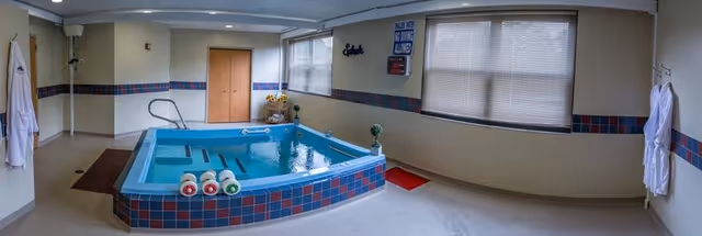 Indoor therapy pool with a tiled surround, handrail, robes on the walls, and windows with blinds.
