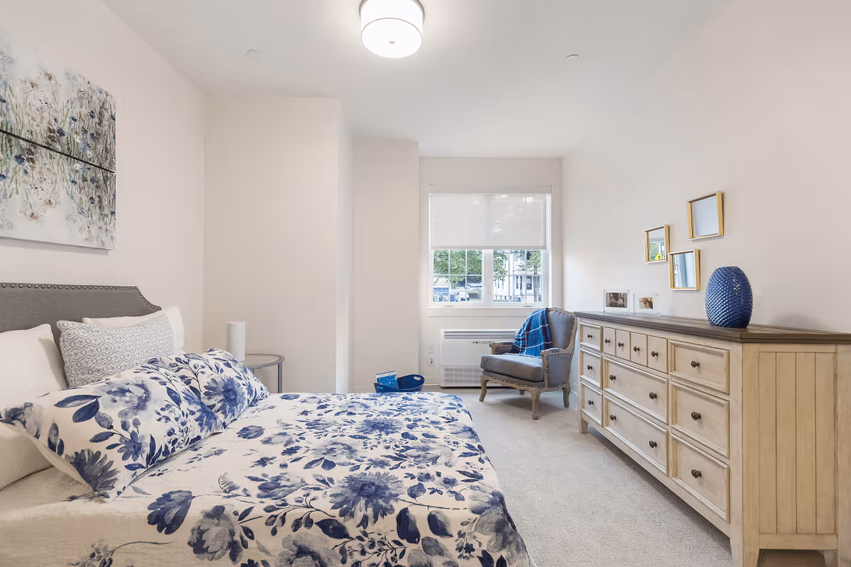 A bright bedroom with a bed covered in a white and blue floral patterned quilt and pillows. There is a gray upholstered headboard, a small round side table with a white lamp, a wooden dresser with multiple drawers, a blue vase, and three small mirrors on the wall above the dresser. A gray armchair with a blue throw blanket is positioned near a window with a white blind, and a white air conditioning unit is below the window.