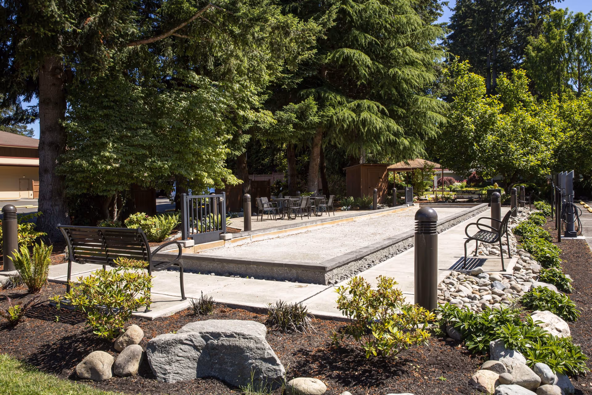 Outdoor garden area with benches, a bocce ball court, trees, shrubs, and a paved walkway under a clear blue sky.