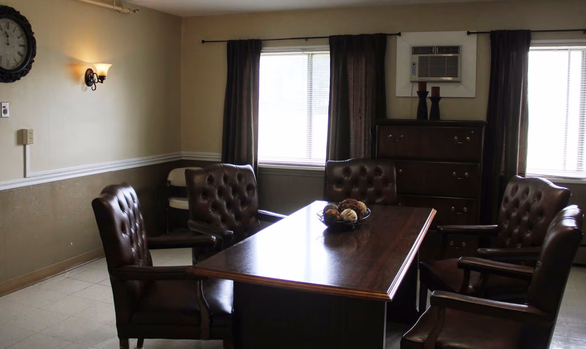 Interior common room with a dark wooden table surrounded by tufted leather chairs, windows with curtains, and a dresser topped by an air conditioner.
