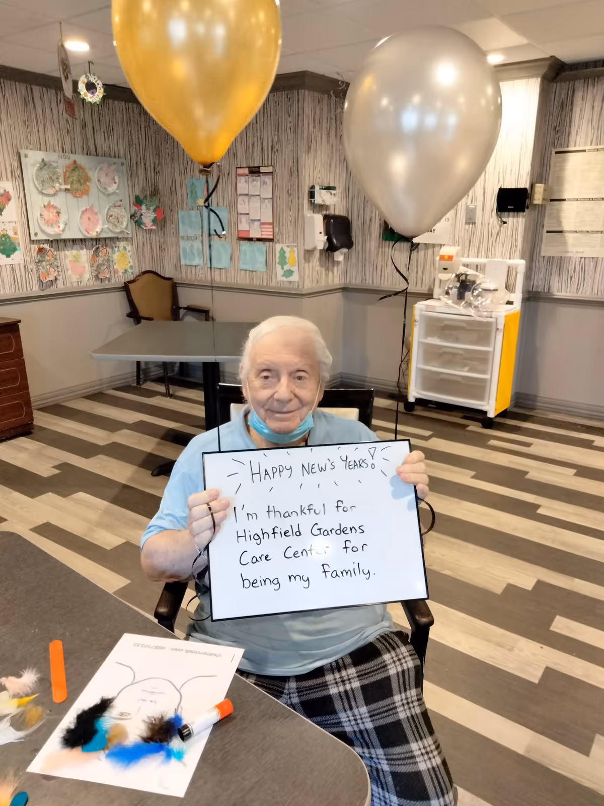 An elderly woman sitting at a table in a decorated room holding a whiteboard sign that reads 'Happy New's Years! I'm thankful for Highfield Gardens Care Center for being my family.' There are two balloons, one gold and one silver, floating behind her. The room has wood-patterned flooring, wall decorations, and a medical cart in the background.